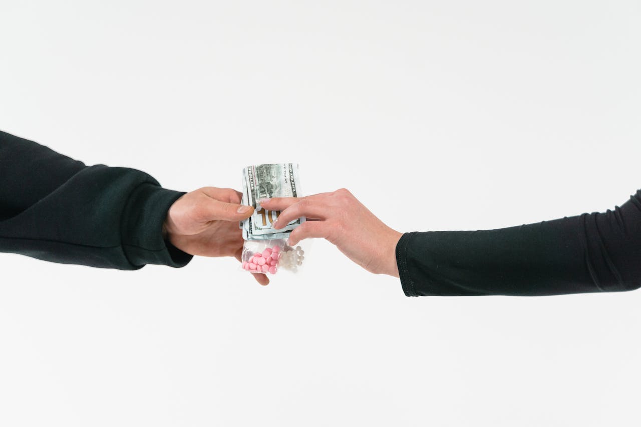 Two hands exchange pills and cash, symbolizing a transaction against a white backdrop.
